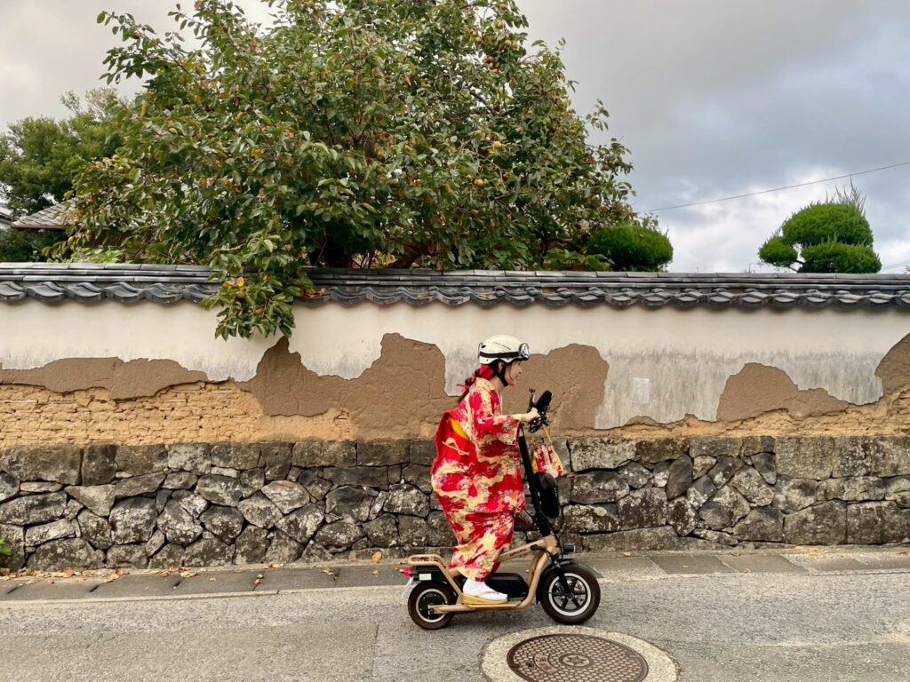 城下町の剥がれかかった趣ある土塀に沿って 着物姿でキックボードに乗る女性